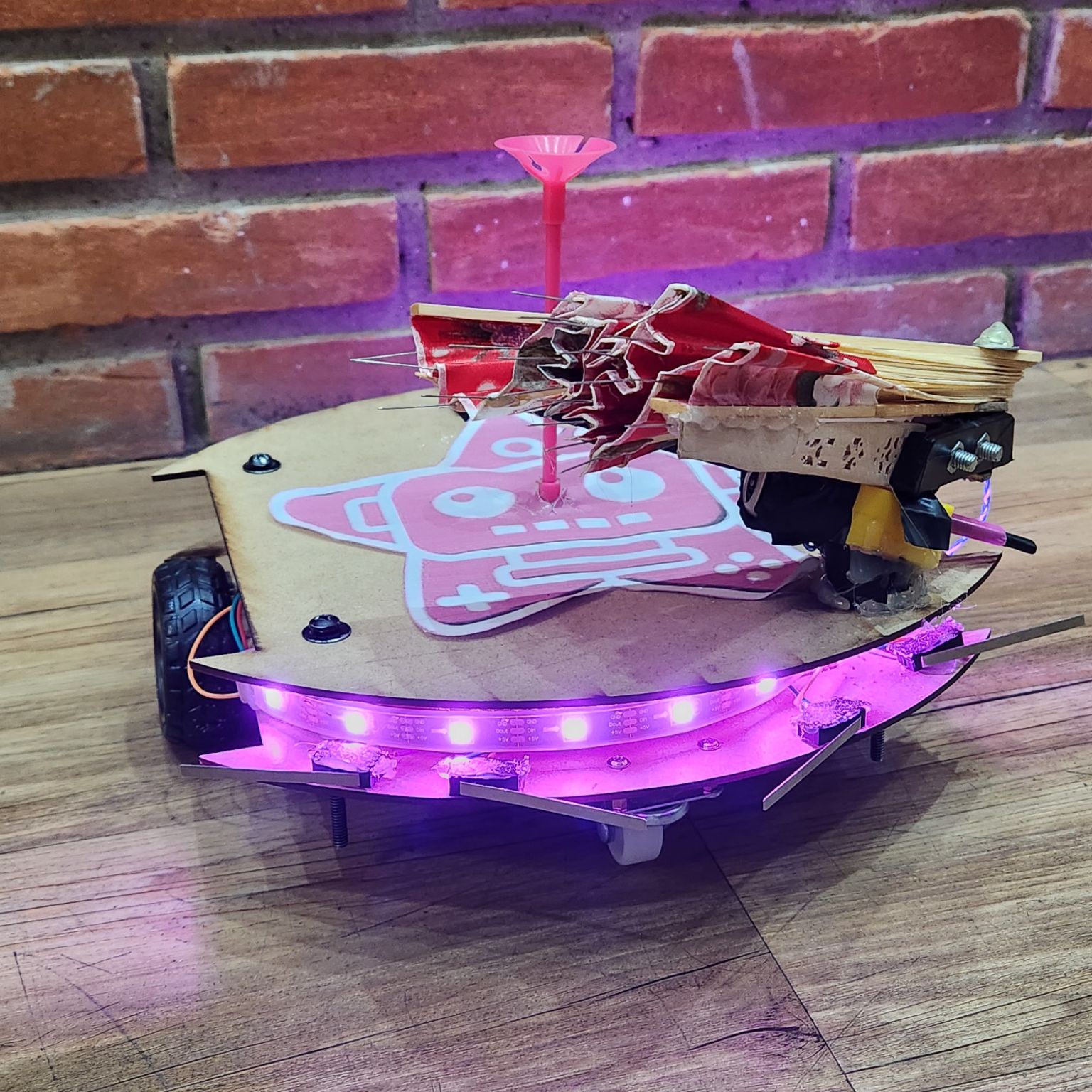 Robô artesanal com base de madeira, LEDs roxos acesos e estrutura de papel colorido, posicionado sobre mesa com fundo de parede de tijolos.